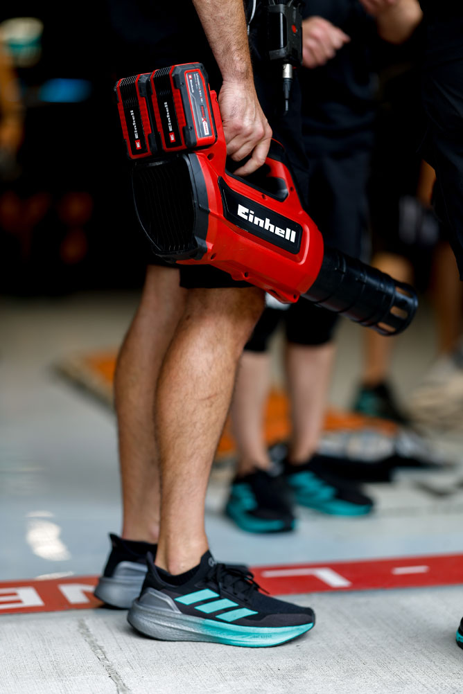 A team member is holding a red Einhell cordless blower with two Power X-Change batteries. The scene takes place in the pit area during a Formula One event.