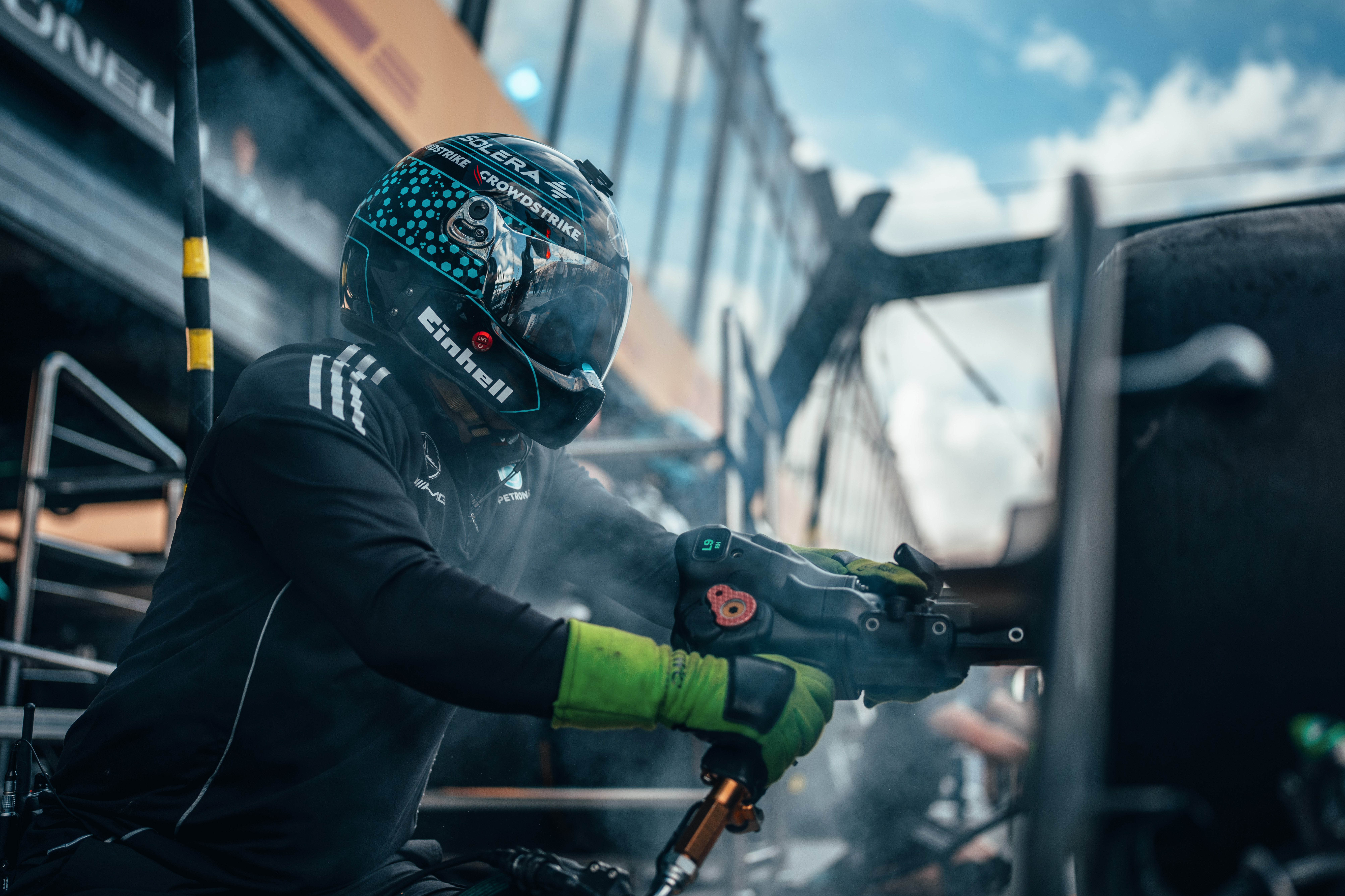 Here’s the translation into English of the sentence you prA member of the Formula 1 pit‑crew wearing a helmet and gloves uses a pneumatic wheel‑gun during a pit stop, surrounded by smoke and equipment.